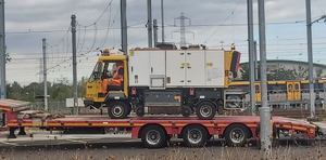 A yellow and white RRV being driven on a low-loader, with a Class 555 unit visible in the background