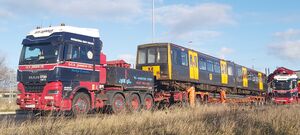 Metrocar 4028 on a red low-loader connected to a red and navy blue truck, with another red and navy blue truck visible alongside it
