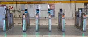 Six metal walls with glass gates attached to the side, forming five gates. All the gates pictured are open. The two outermost gates are wider than the middle three. Every wall has two light strips on the front; all the strips pictured are either off or lit green. Every wall has a computer screen with instructions and an LED arrow pointing to the corresponding gate, both on the top. Every wall except the leftmost one is fitted with an orange paper ticket feeder and a black NFC/contactless reader, both at the top front corner.