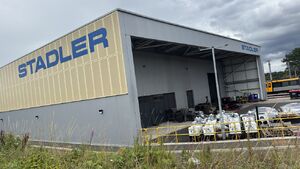 Image shows a modern building with a grey wall and yellow-creme coloured panels above door height. On the panels there is a blue Stadler logo. There is a smaller blue stsdler logo on the other side of the building, above a storage shed.