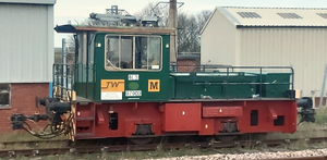 A locomotive numbered BL3 and 97903, with a green body, red undercarriage, and a plaque, alongside two buildings