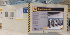 Cream-coloured walls enclosing a construction area. A locked entrance with windows is visible on one side, and an information panel labelled "Gateline project" and branded with the NECA logo on the other.